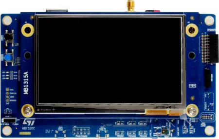 The image displays a blue electronic board featuring a central LCD screen area. Various connectors, buttons, and labeled components are visible on the edges. Notably, it includes labels such as "MB315A" and "MB1520C," suggesting it may be part of a development or display module.