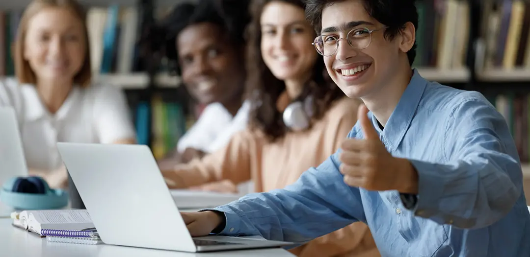 Student in front of laptop, giving a thumbs up, several students in background